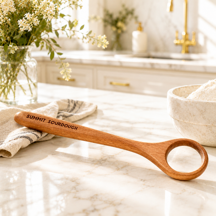 Premium wooden Danish dough whisk engraved with Summit Sourdough resting on a bright marble countertop in a luxury farmhouse kitchen beside a ceramic flour bowl and soft linen cloth, to make sourdough bread.