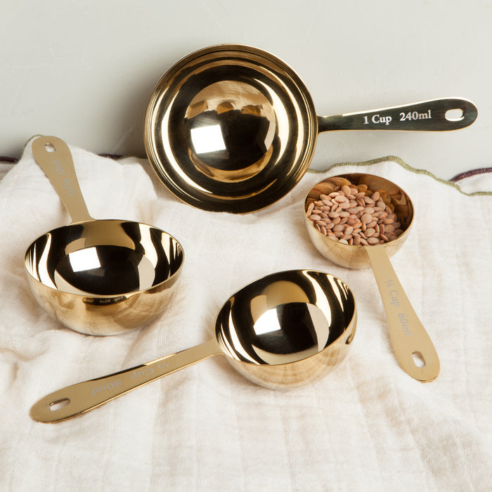 Danica Heirloom gold measuring cups displayed with baking ingredients on a wooden countertop.
