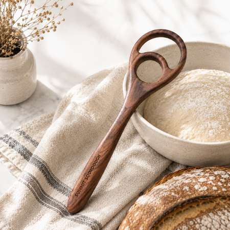 Handcrafted double loop sourdough dough whisk made from wood resting on a marble counter beside flour and linen in a premium baking kitchen.