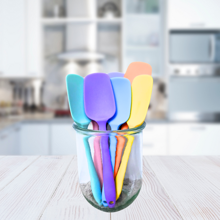 A collection of silicone spatulas in various solid colors displayed inside a clear glass on a kitchen counter.